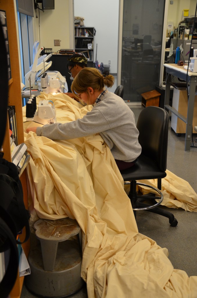 Student sits at a sewing machine, sewing a huge amount of beige fabric