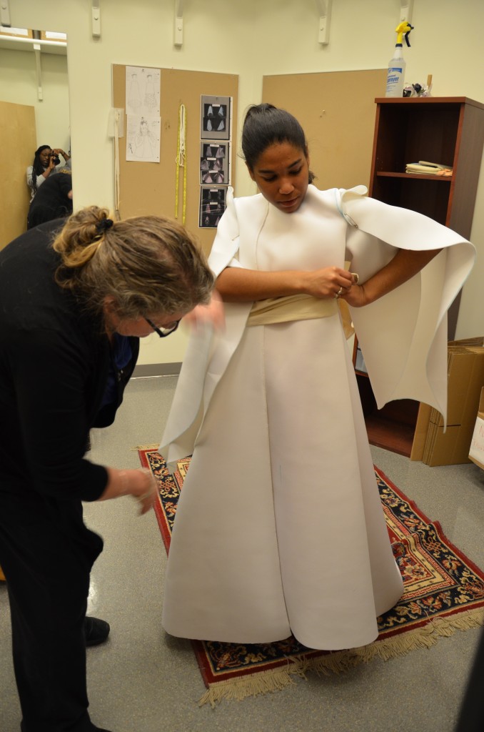 Large pieces of white fabric are pinned onto a student to form a dress in a fitting