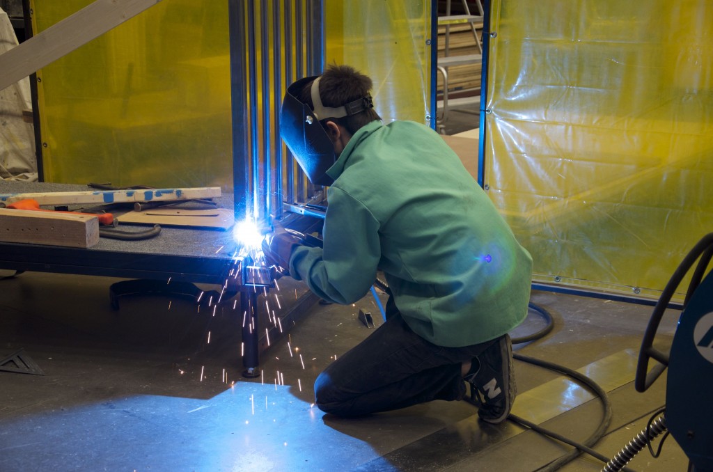 Student welds metal, wearing a helmet, gloves, and jacket.