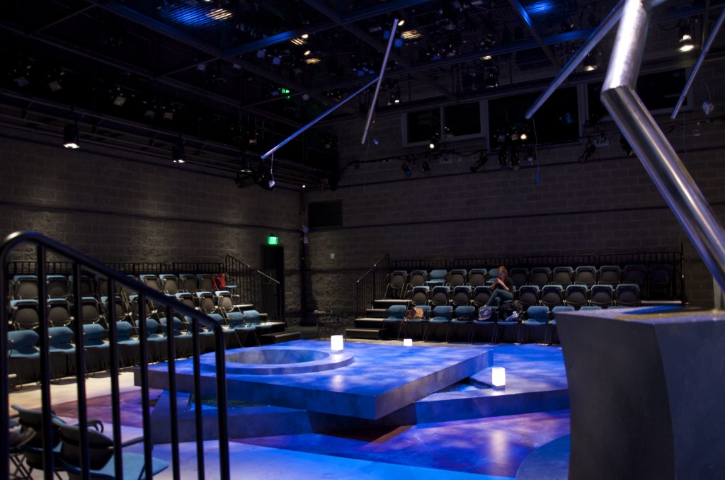 Set in the Black Box theatre, lit up with blue and purple lights. Set consists of two large, grey platforms skewed on a 45 degree angle.