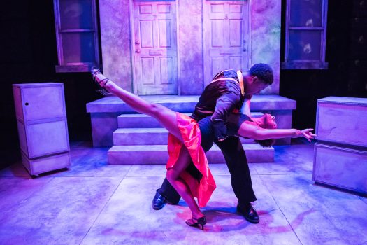 One male-presenting actor dips a female-presenting actor in a dance. She is wearing a red skirt and black top, and sticking her leg out. He wears all black. In the background is a set of stairs, leading to two doors and a wall, which look purple and blue under the lights.