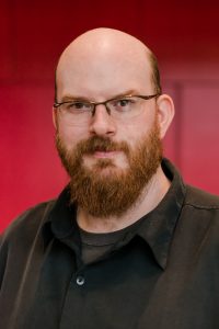 Evan McDougall, a white man with minimal hair on his head, a red beard, and rectangular glasses
