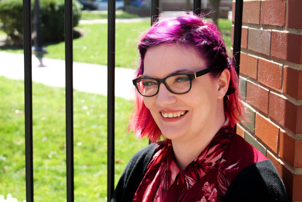 Headshot of Margaret Caster. She is white, with pink hair and glasses.